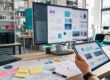 KI Tools für Unternehmen: Arbeitsraum einer Marketingagentur mit Technik, Farbmuster und Servern, männliche Hand mit Tablet.